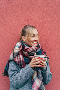 Smiling young woman using mobile phone while standing against red wall