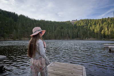 Woman wearing hat while standing on pier by lake against sky