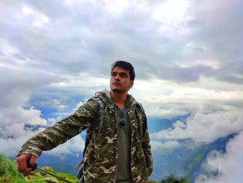 Young man looking away while standing on mountain against cloudy sky