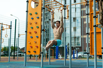 Shirtless young man exercising on gymnastic rings against building