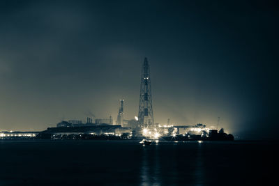 Illuminated factory by sea against sky at night