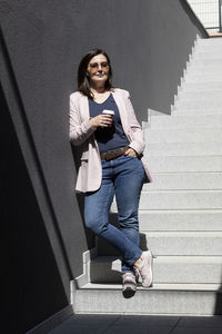 Smiling beautiful brunette woman hold paper cup of coffee standing on stairs near office outdoor