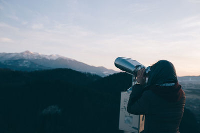 Woman looking at mountains through coin-operated binoculars against sky
