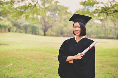 Portrait of young woman in graduation gown holding certificate while standing at park