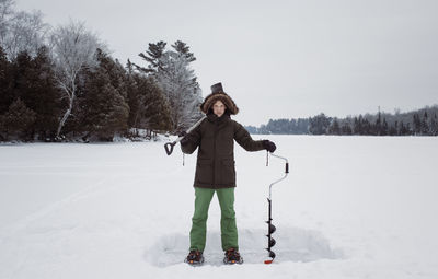 Portrait of man holding ice auger and shovel while standing on frozen lake against sky