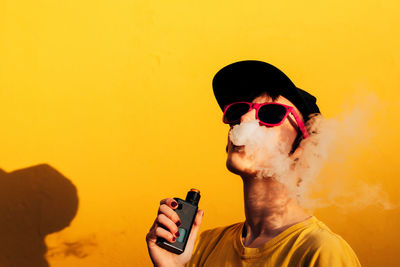 Contemporary female in stylish outfit exhaling fume while standing near yellow wall and vaping on city street