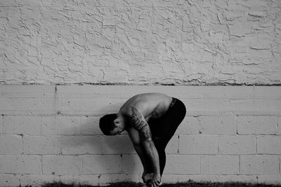 Side view of shirtless man tying shoelace against wall