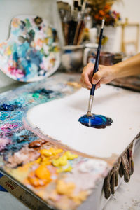 Crop view of hand of talented female artisan taking paint with brush while creating picture on canvas in art workshop