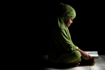Girl wearing hijab reading koran while sitting on floor against black background