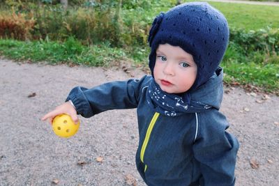 Cute baby girl holding ball while standing on dirt road by grassy field