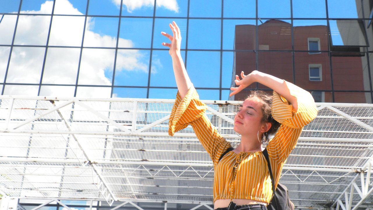 YOUNG WOMAN WITH ARMS RAISED STANDING AGAINST BUILDING