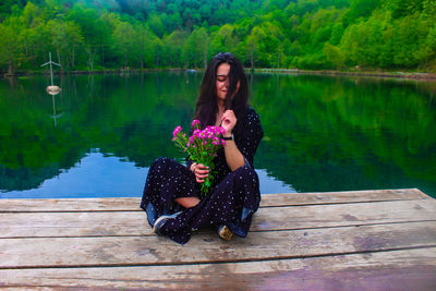 Full length of young woman holding flowers while sitting on pier over lake