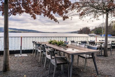 Empty chairs and tables by lake against sky