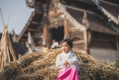 Girl looking away sitting on haystack against barn