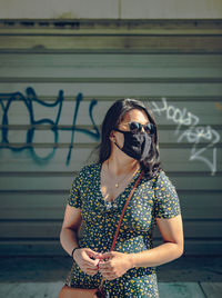 Young woman wearing sunglasses and mask while standing against wall