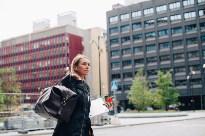 Mature businesswoman walking with luggage against buildings in city