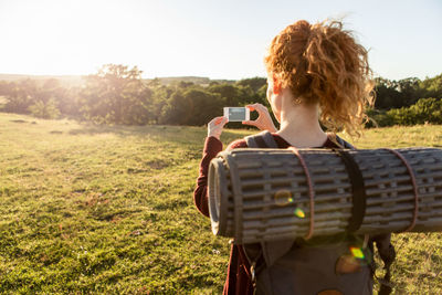 Rear view of woman with backpack photographing field against sky during sunset