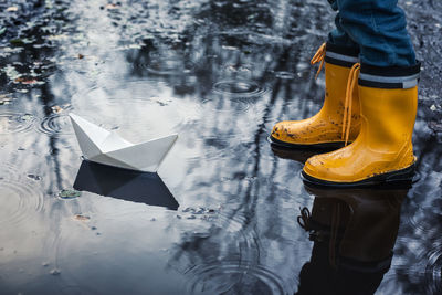 Low section of boy standing by paper boat in puddle