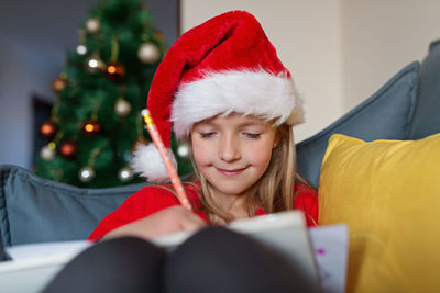 Cute blonde caucasian girl 7 years old sitting on couch near christmas tree writing letter to santa