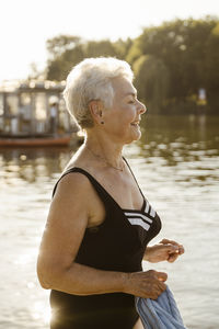 Smiling elderly woman wearing swimsuit while standing near lake on vacation