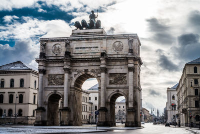 Low angle view of siegestor with buildings against cloudy sky