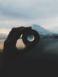 Cropped hand holding camera lens in front of river and mountain