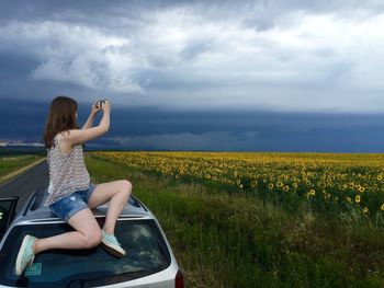 Woman sitting on car roof while photographing sunflower field against cloudy sky