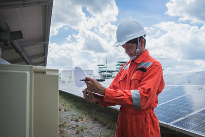 Side view of worker wearing reflective clothing while standing on solar panel against sky