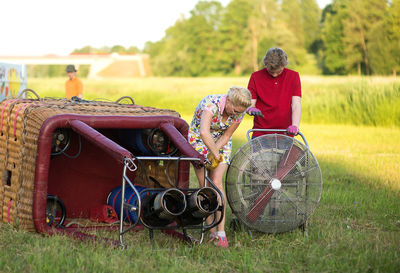Boy assisting pilot in preparing hot air balloon on grassy field during sunset