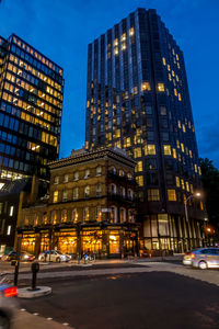 Illuminated street by buildings against sky at night