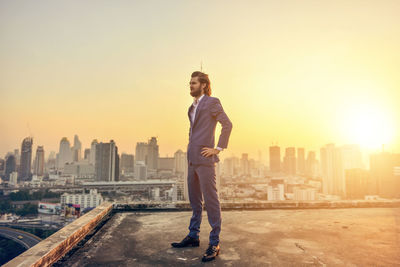 Businessman looking at cityscape while standing on building terrace against clear sky during sunset