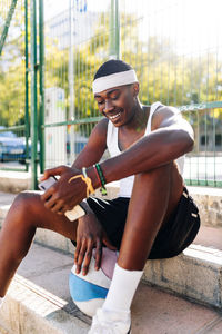 Happy african american male basketball player sitting on sports ground and browsing smartphone on sunny day in summer