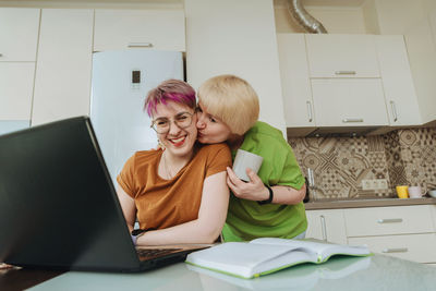 An elderly mother kisses her daughter, who is using a laptop