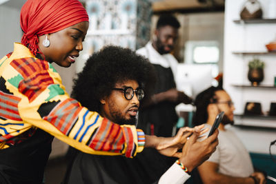 Female hairdresser using smart phone of male customer in barber shop