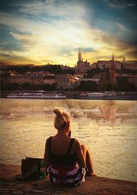 Rear view of woman sitting on riverbank against sky during sunset