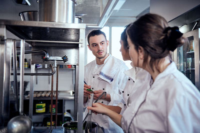 Male chef taking notes while looking at colleagues cooking in commercial kitchen