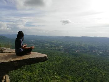 Woman meditating while sitting on rock against sky