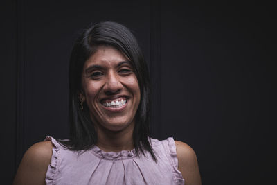 Positive self assured young latino female wearing casual blouse smiling and looking at camera while standing against black background