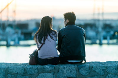 Rear view of couple sitting on retaining wall by river