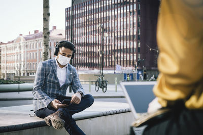 Businessman wearing face mask sitting outside using smartphone