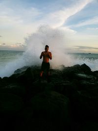 Shirtless man standing against splashing wave at beach