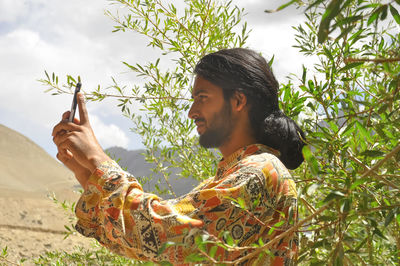 Side view of a long haired young guy taking photo with his phone with the background of mountains 