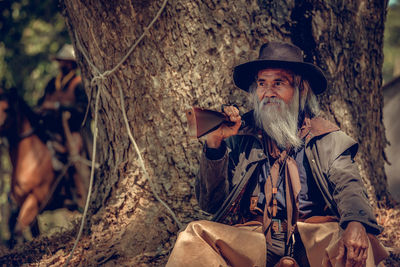 Bearded senior man holding gun while sitting by tree