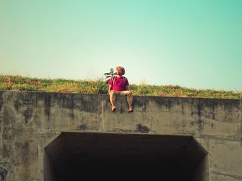 Full length of man sitting on retaining wall against clear sky