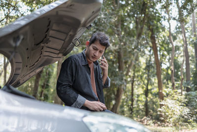 Man talking on while standing by broken vehicle against trees