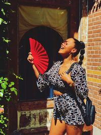 Smiling young woman holding hand fan while standing against brick wall