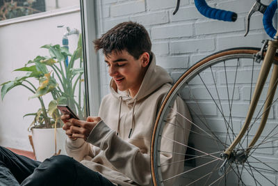 Young man using mobile phone while sitting against wall outdoors