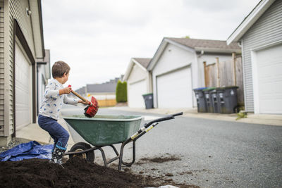 Young boy scooping pile of soil into wheelbarrow in back alley