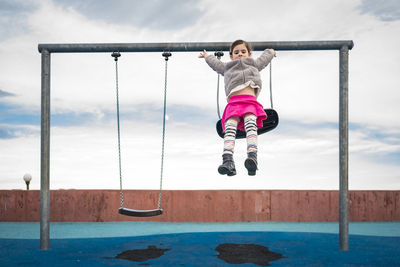 Playful girl jumping on sand from swing against cloudy sky at playground