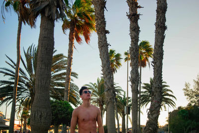 Shirtless young man standing against trees during sunset